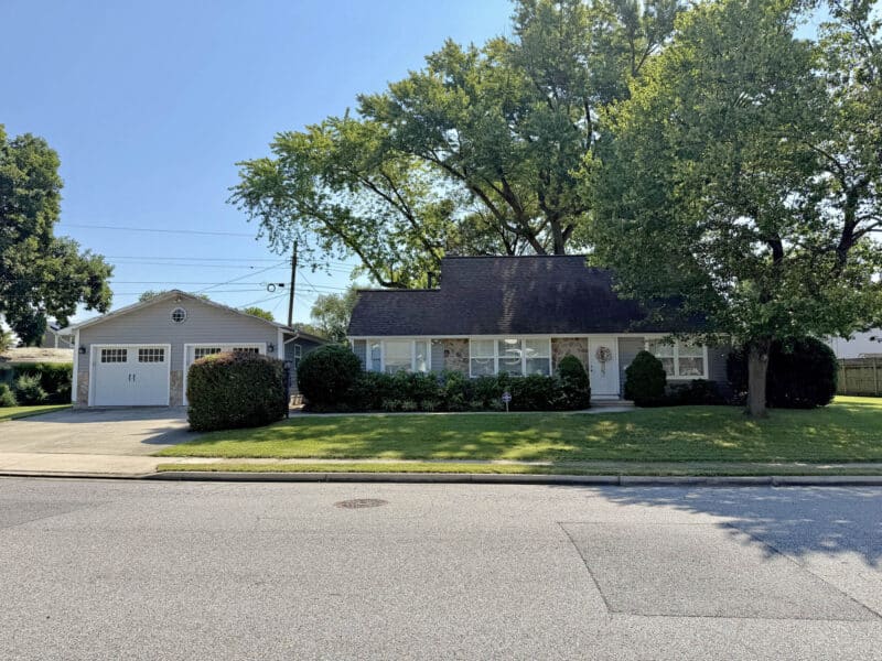 Wide front view of Cape Cod home with detached two-car garage, driveway, mature trees and landscaped lawn.