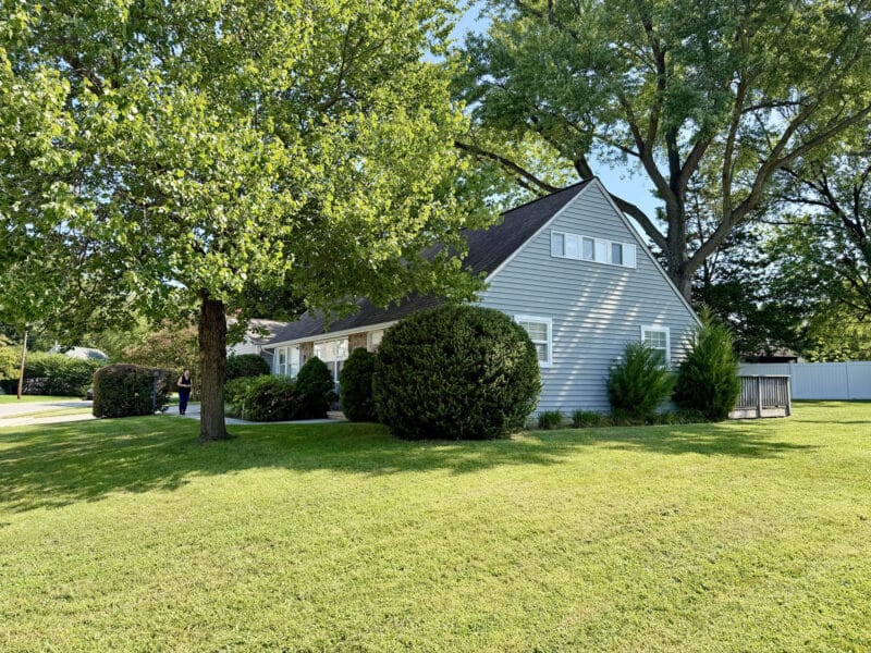 Side exterior of Cape Cod home with siding, mature landscaping, large lawn and rear deck.