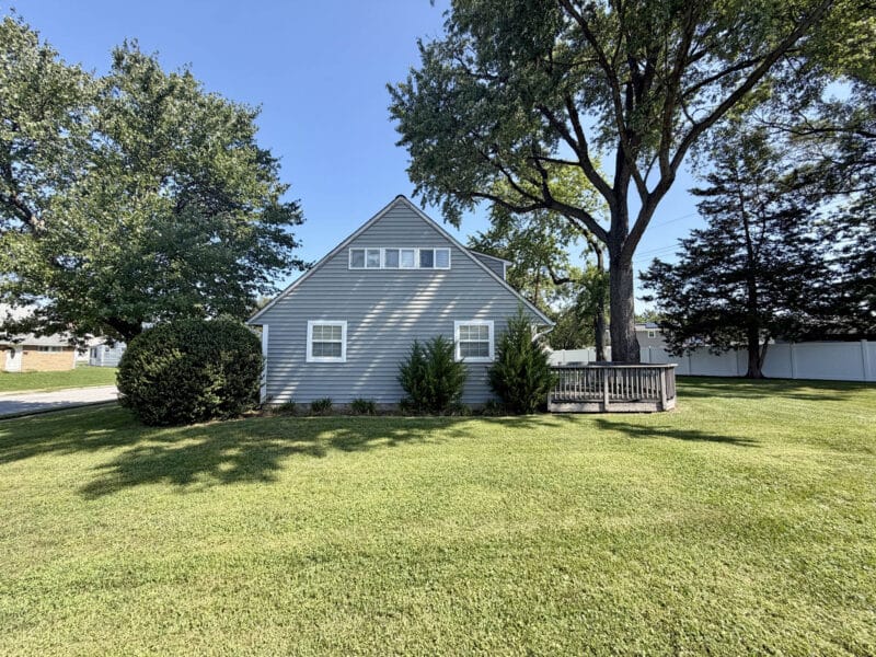 Side exterior of Cape Cod home with large lawn, mature shade trees and wood deck.