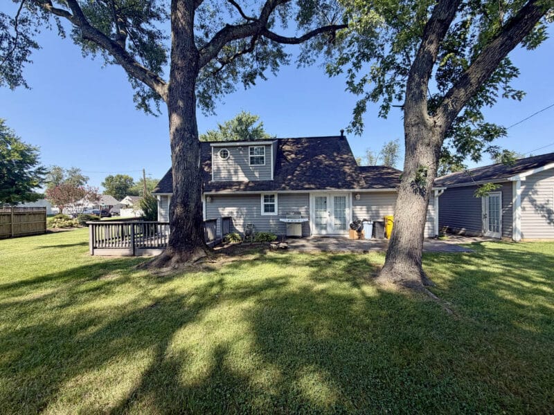 Rear exterior of Cape Cod home with patio, small deck, mature trees and landscaped backyard.