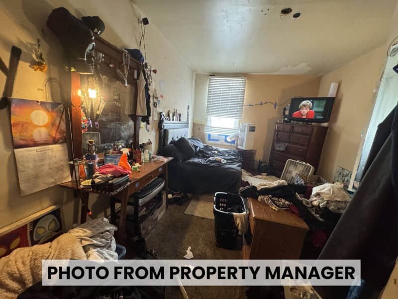 Property manager photo of a furnished bedroom at 4900 Curtis Avenue showing bed, dresser, window air conditioner, and personal belongings.