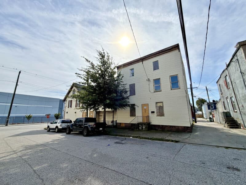 Rear exterior of 4900 Curtis Avenue in Curtis Bay, Baltimore, showing vinyl siding, multiple entrances, boarded windows, and adjacent alley.