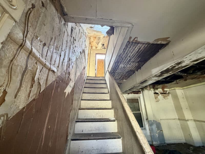 Interior wooden staircase at 4900 Curtis Avenue in Baltimore with peeling wall finishes, damaged plaster, and exposed ceiling lath.